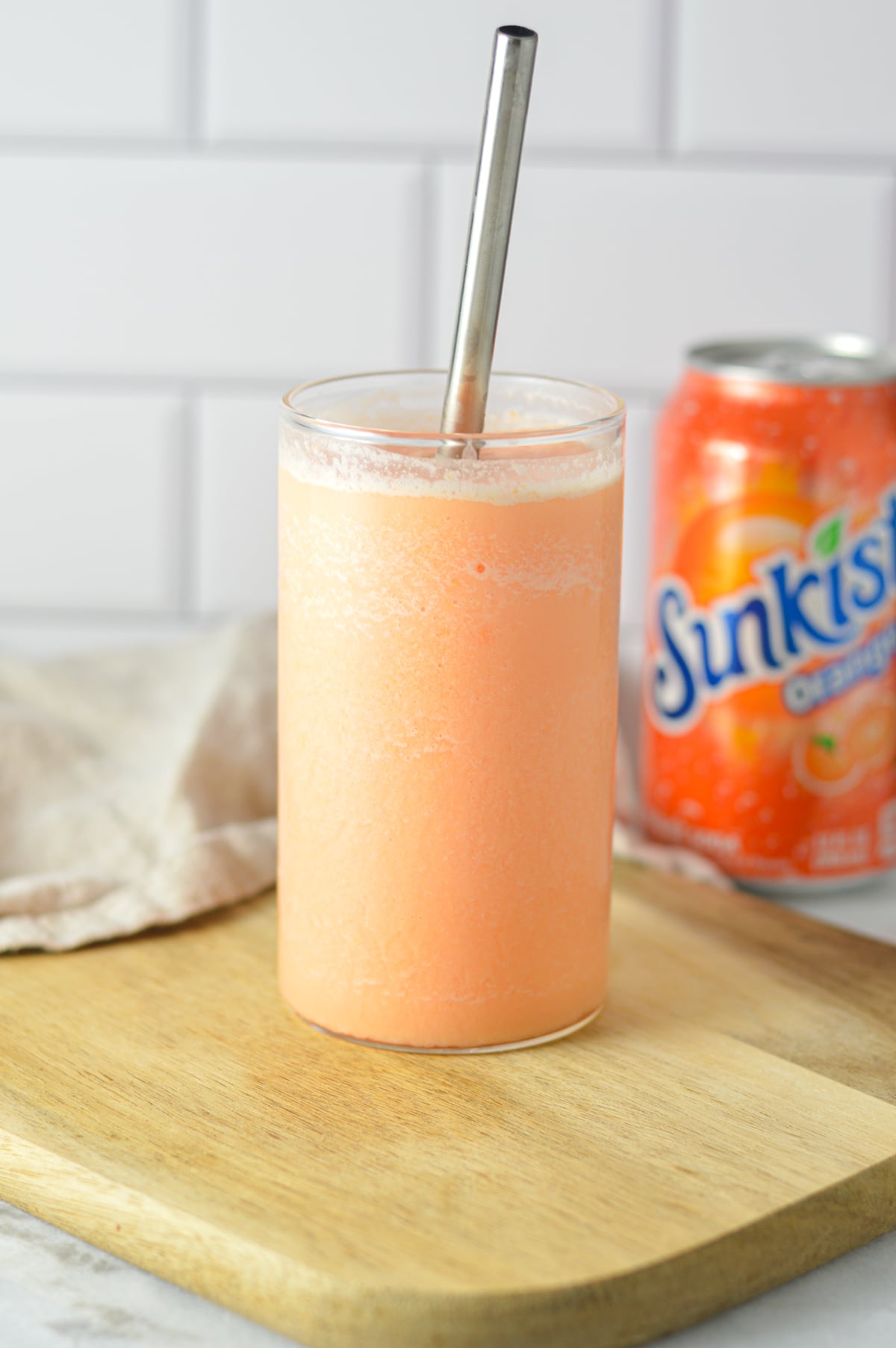 A tall glass filled with a slushy orange creamsicle drink, next to a can of Sunkist Orange soda on a kitchen counter.