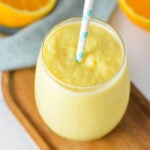 A glass of creamy orange slushie with a blue pattern straw served on a wooden surface with fresh orange slices in the background.