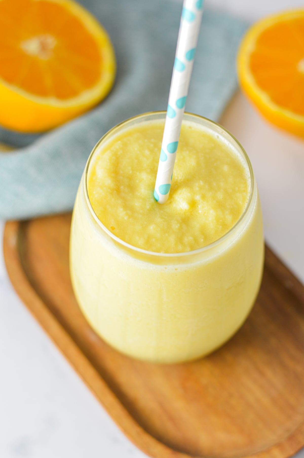 A glass of creamy orange slushie with a blue pattern straw served on a wooden surface with fresh orange slices in the background.