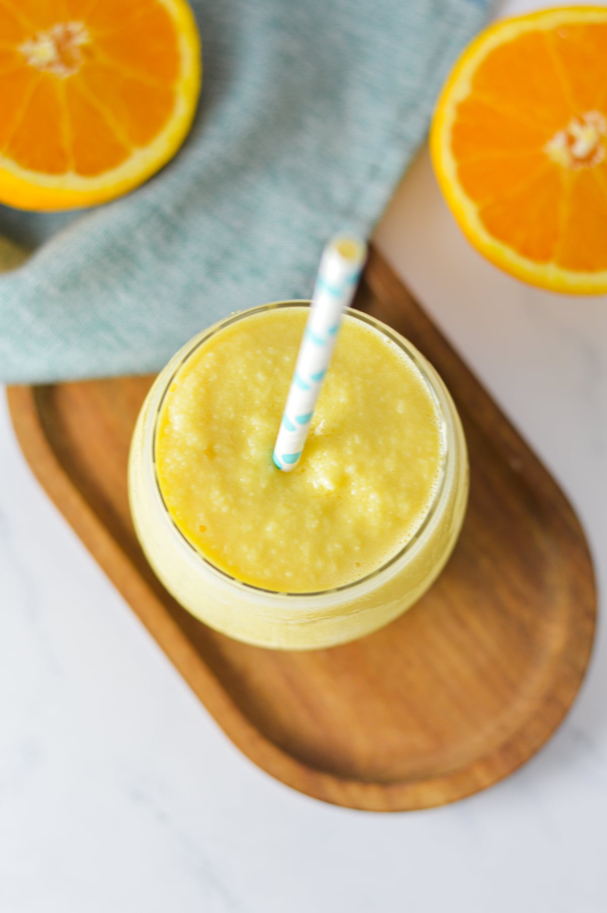 A top-down view of a glass of creamy orange Julius slushie with a blue pattern straw, placed on a wooden board and fresh orange slices in the background.