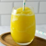 A glass filled with a slushy yellow orange mimosa drink, topped with a light blue patterned straw on a wooden coaster.