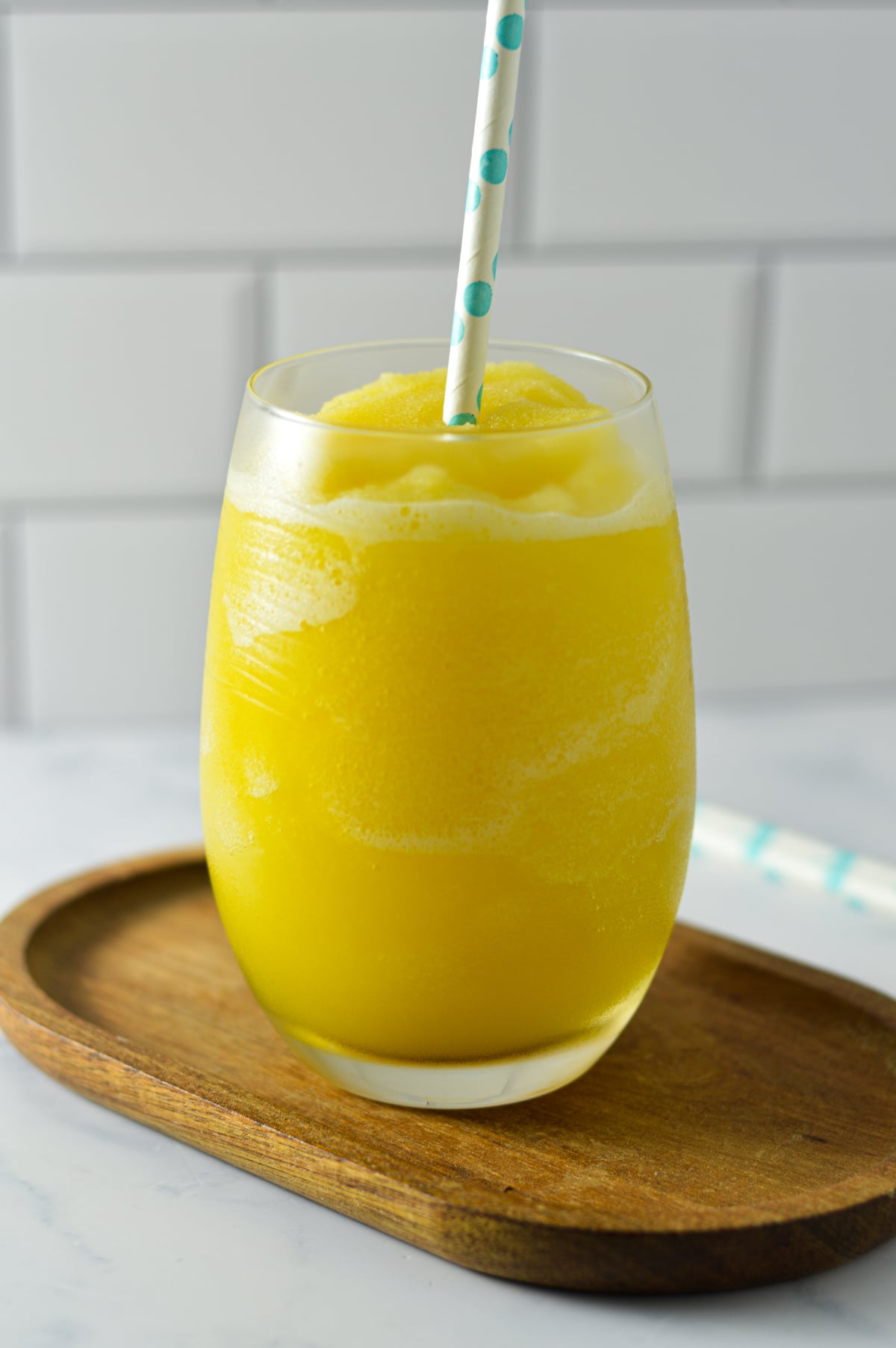 A glass filled with a slushy yellow orange mimosa drink, topped with a light blue patterned straw on a wooden coaster.