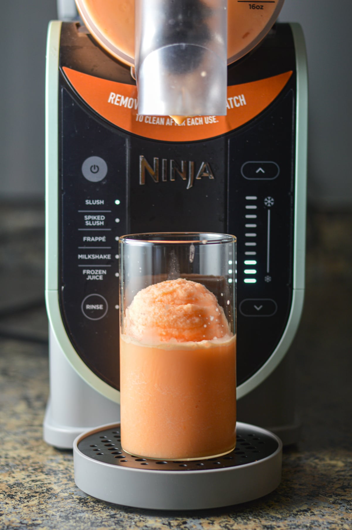 A clear plastic cup of slushy orange creamsicle drink being filled directly from the dispenser spout of a Ninja Slushi machine.