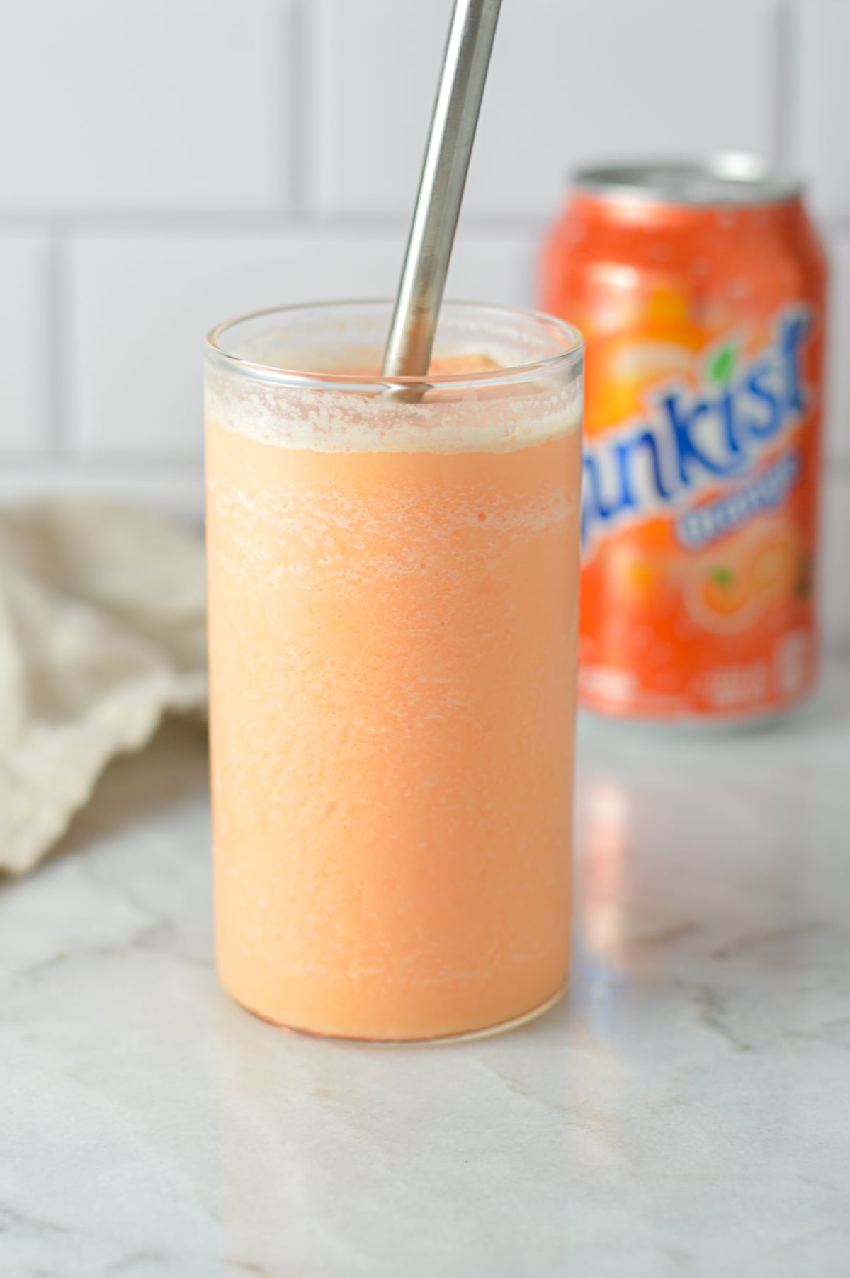 A close-up of a tall glass filled with a slushy orange creamsicle drink, positioned in front of an orange Sunkist soda can.