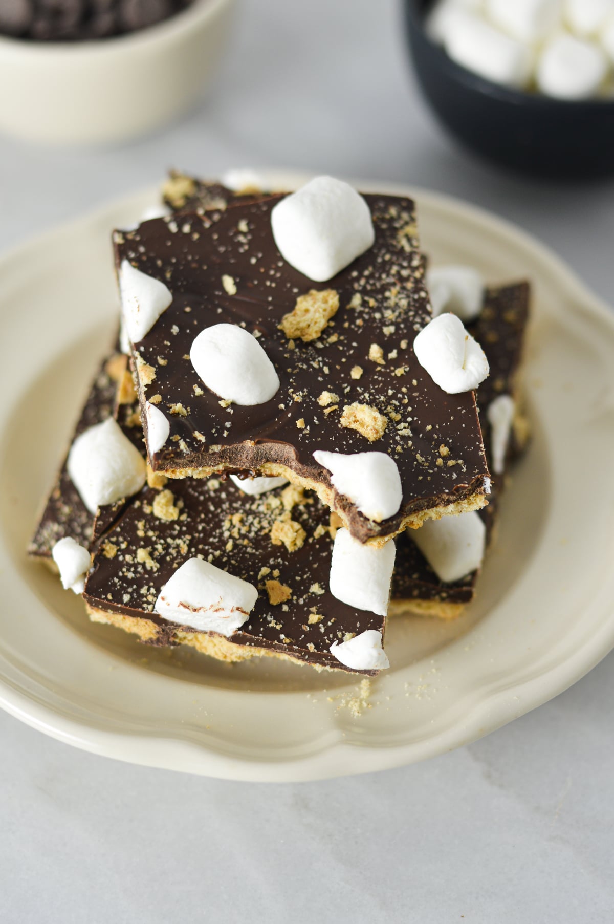 A stack of S'mores Chocolate Bark squares layered over graham crackers and topped with mini marshmallows and crushed graham cracker crumbs.