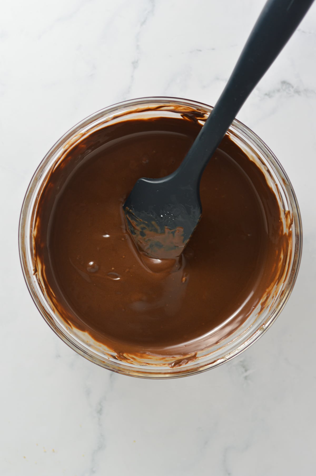 A clear bowl filled with melted chocolate with a rubber spatula mixing the chocolate.