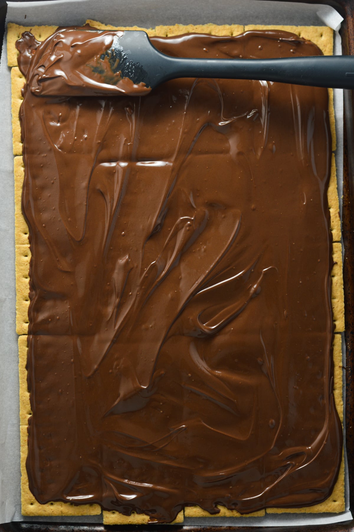 Melted chocolate spread evenly over a single layer of graham crackers on a parchment-lined baking sheet.